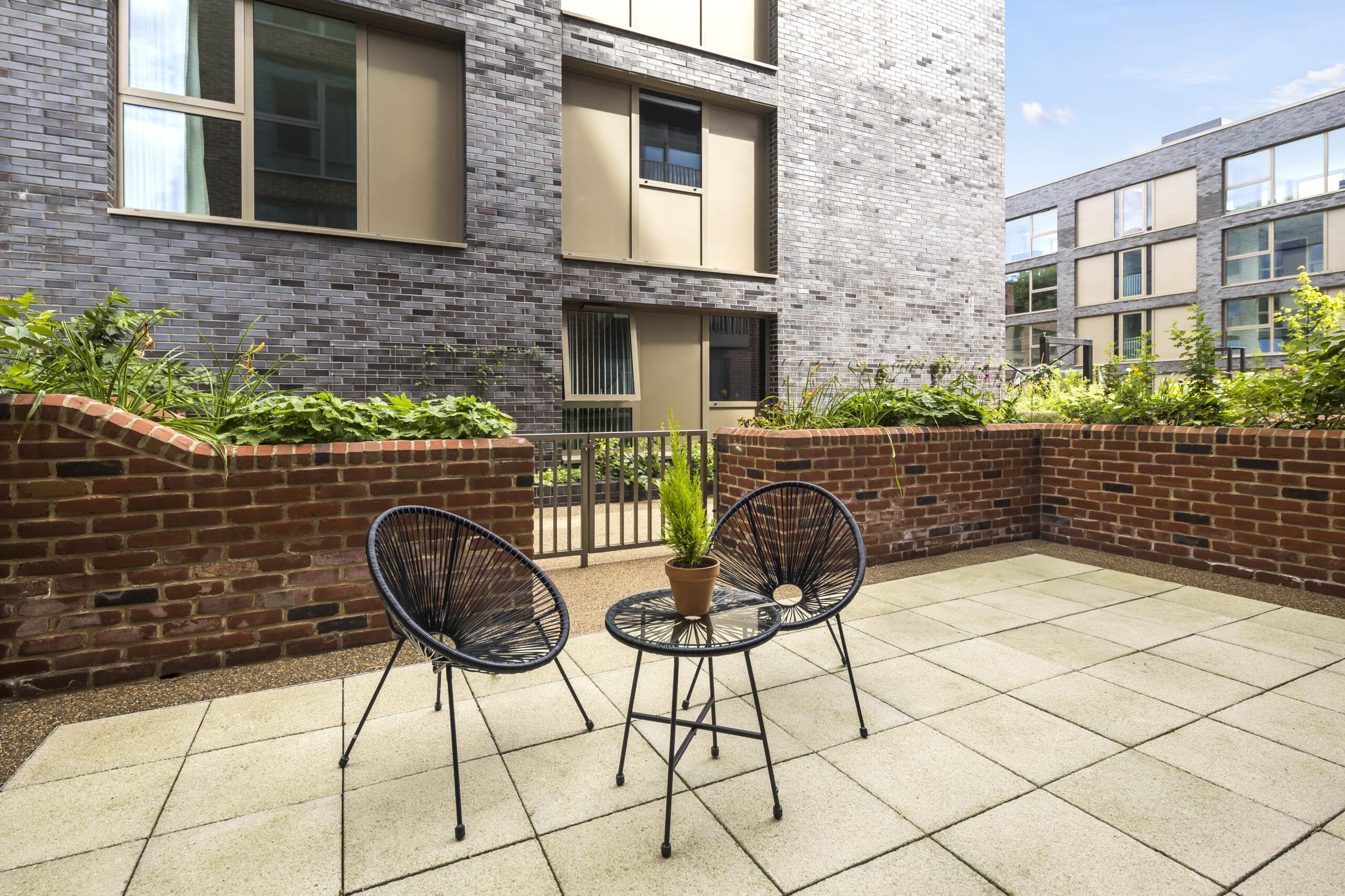 A private outdoor terrace with 2 garden chairs and a small table. It is surrounded by a brick wall that includes planters full of greenery.