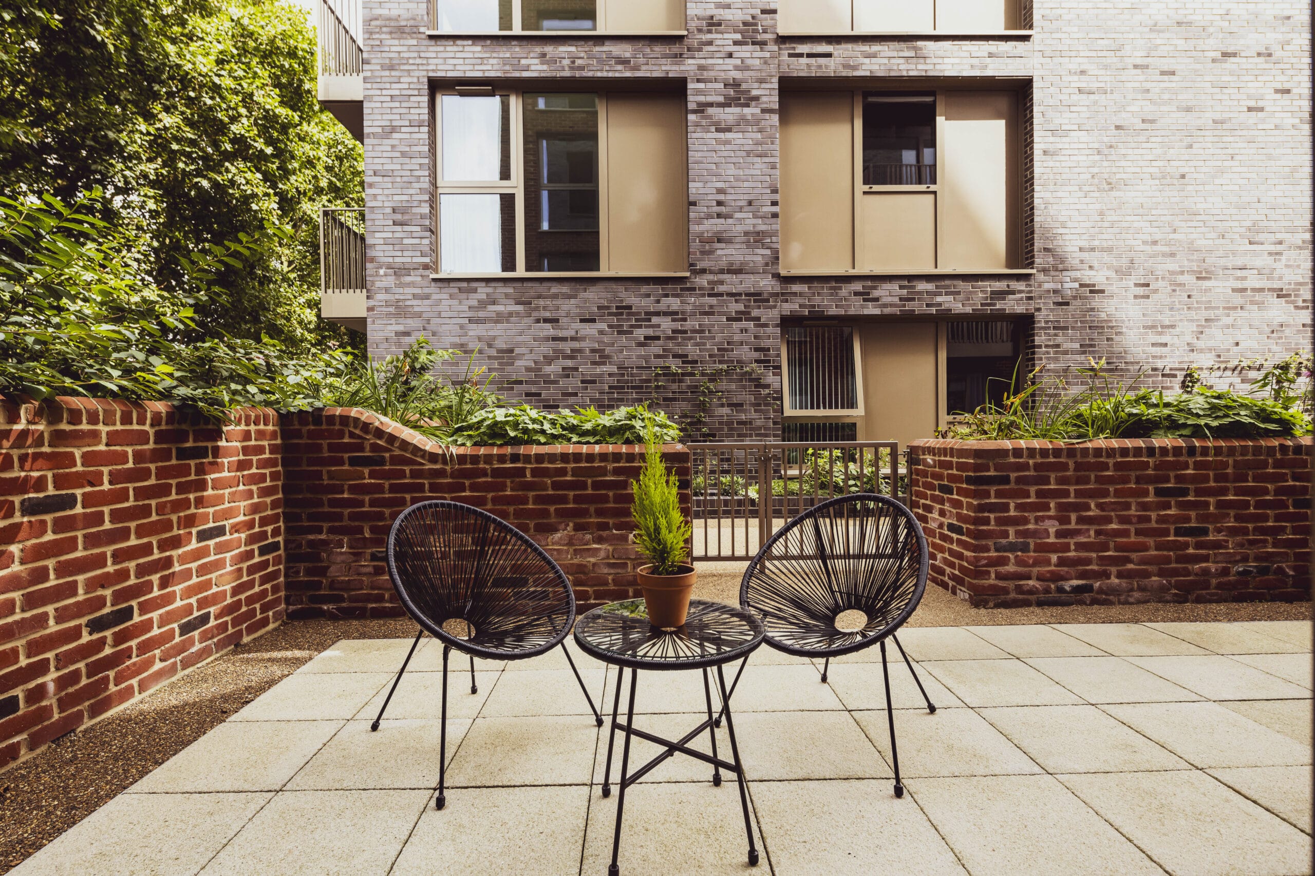 A private outdoor terrace with 2 garden chairs and a small table. It is surrounded by a brick wall that includes planters full of greenery.