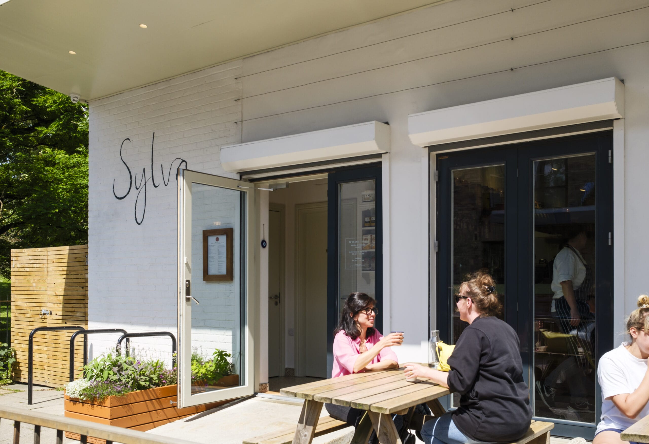 An outdoor shot of the entrance to Sylva Cafe in Deptford. There are 3 people using the outdoor seating.