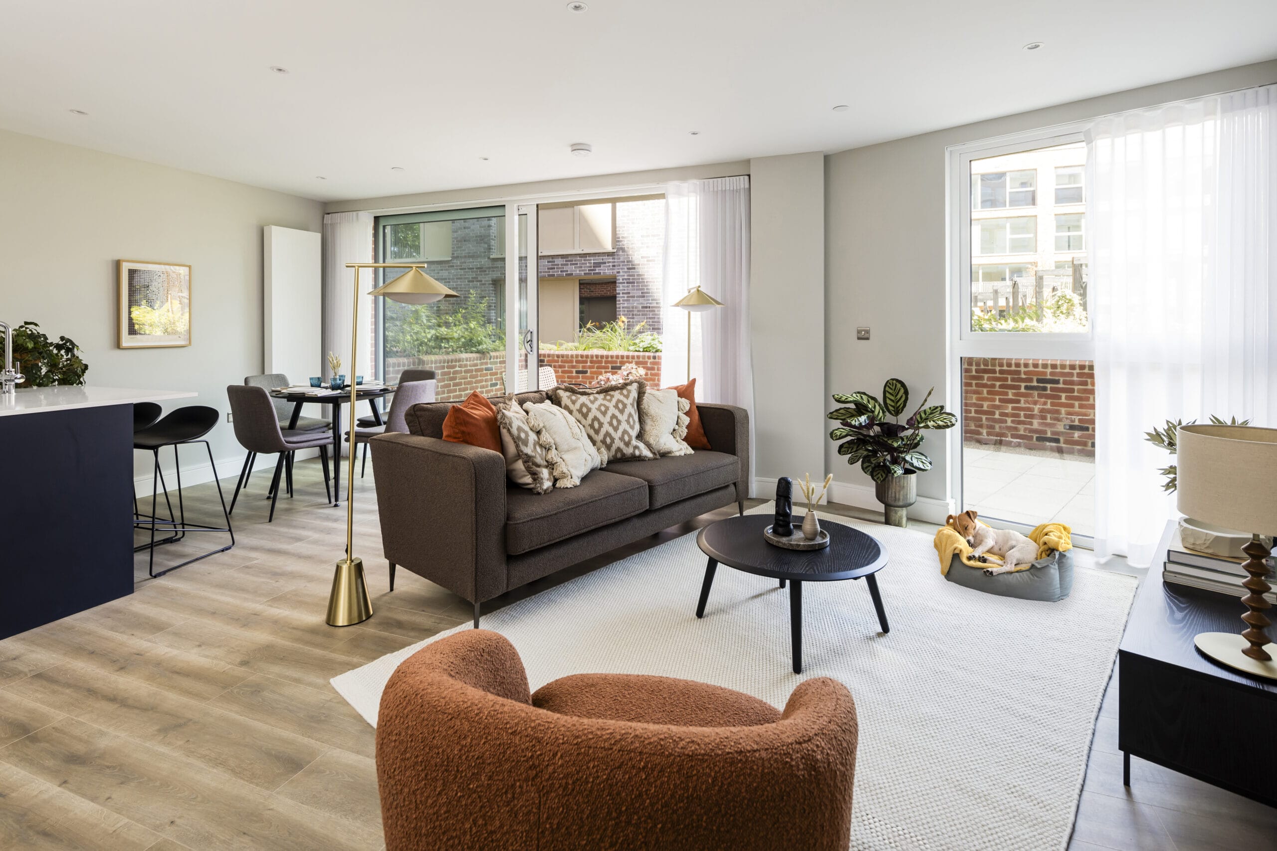 A modern spacious open plan living/dining room with a sofa, armchair, coffee table, dining table and floor lamps. The balcony is visible in the background of the image. There is a small dog sleeping in front of the window - showing that this apartment is pet friendly.