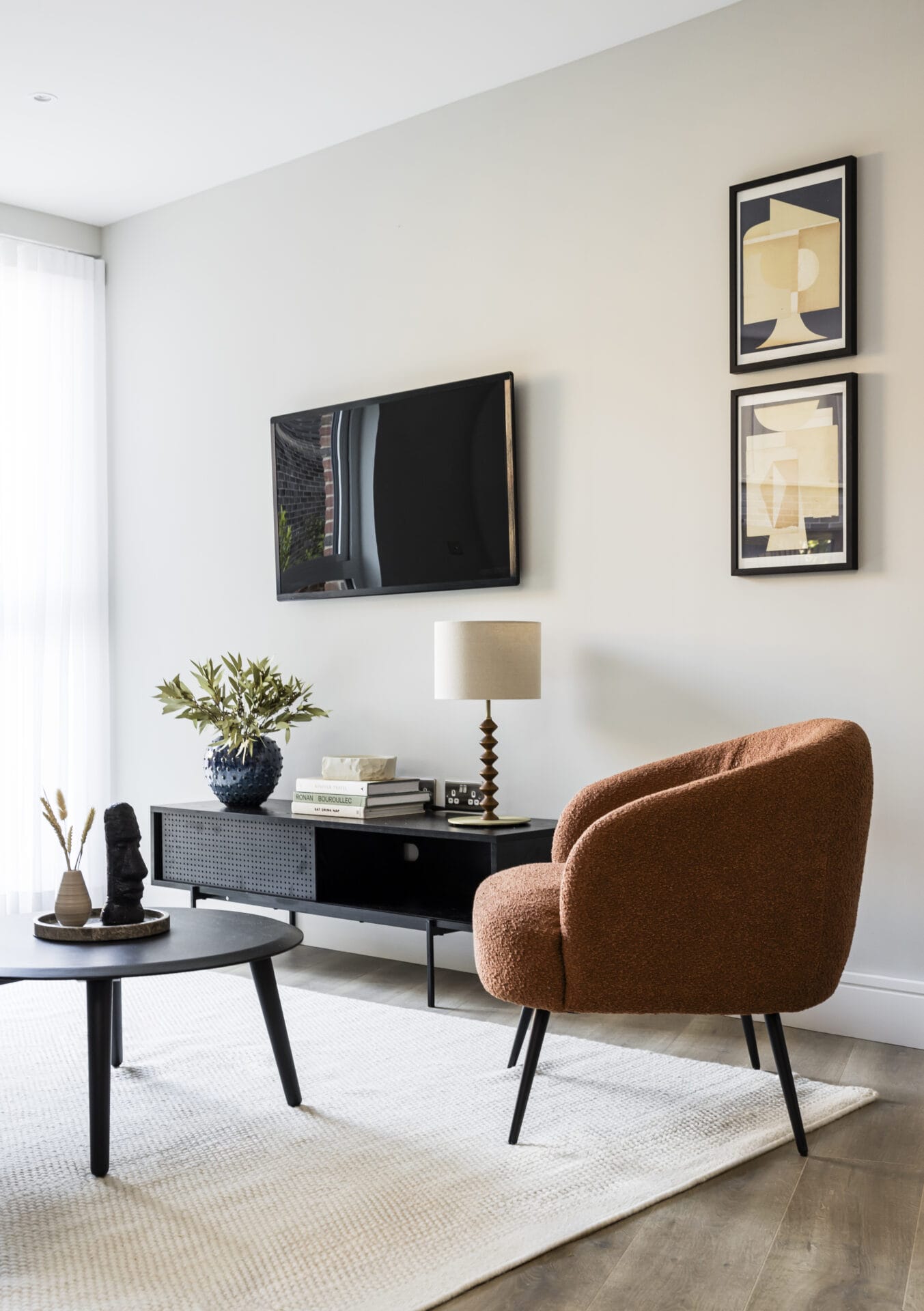 An interior of the living room with an armchair, a TV mounted on the wall, a coffee table and various items of decor such as a lamp, vase and framed art.