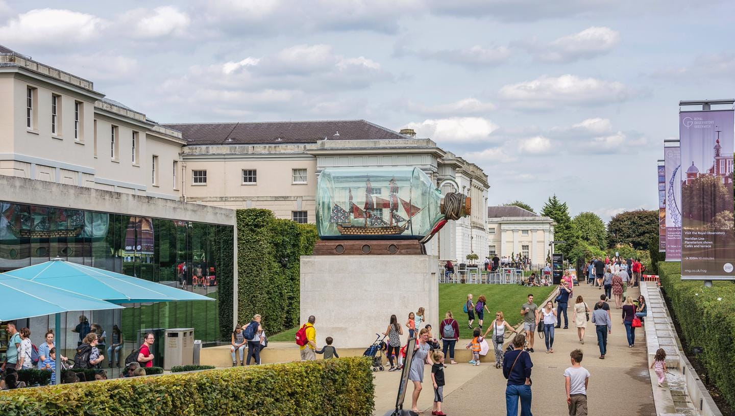 The grounds of the National Maritime Museum. There are lots of people enjoying the outdoor areas and a large sculpture of a ship in a bottle sat in the centre.