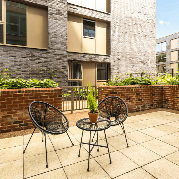 A private outdoor terrace with 2 garden chairs and a small table. It is surrounded by a brick wall that includes planters full of greenery.